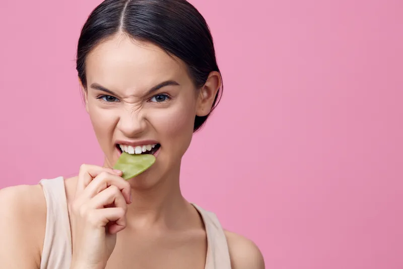 Young woman playfully biting leaf with a mischievous expression against a pink background, showcasing a playful attitude towards healthy eating