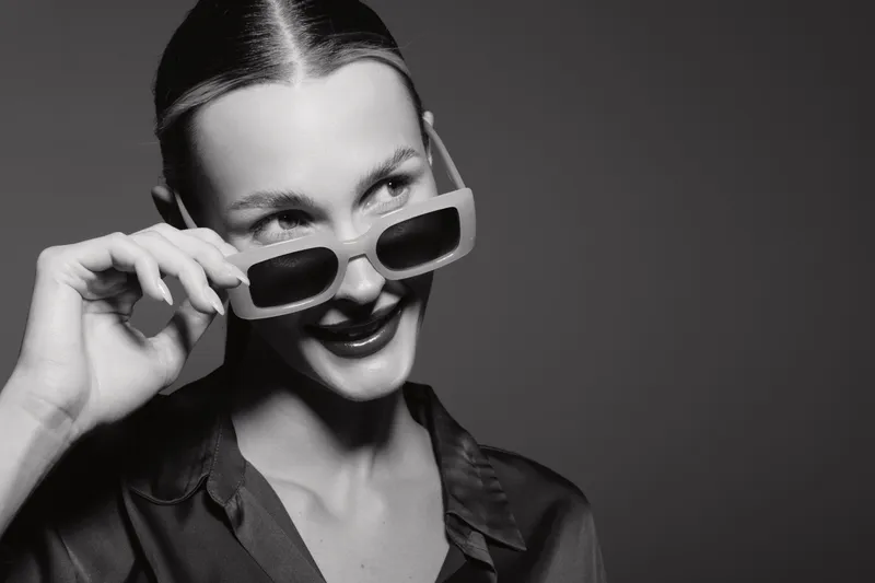 Stylish white woman with pale skin wearing sunglasses and silk shirt looks sideways with a smile on dark background. Concept of fashion, elegance, and confident female model.