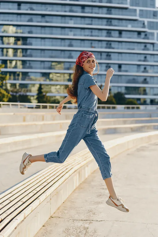 Lifestyle woman in modern boho-western denim street style jumps outdoors, wearing a red bandana and casual shoes with vibrant film color tones and urban background.