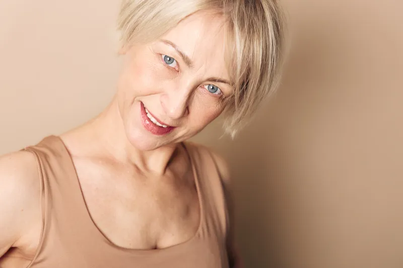Smiling middle aged woman with short blonde hair and blue eyes wearing beige tank top against light beige background. Concept of natural beauty, confidence, and positive mood.