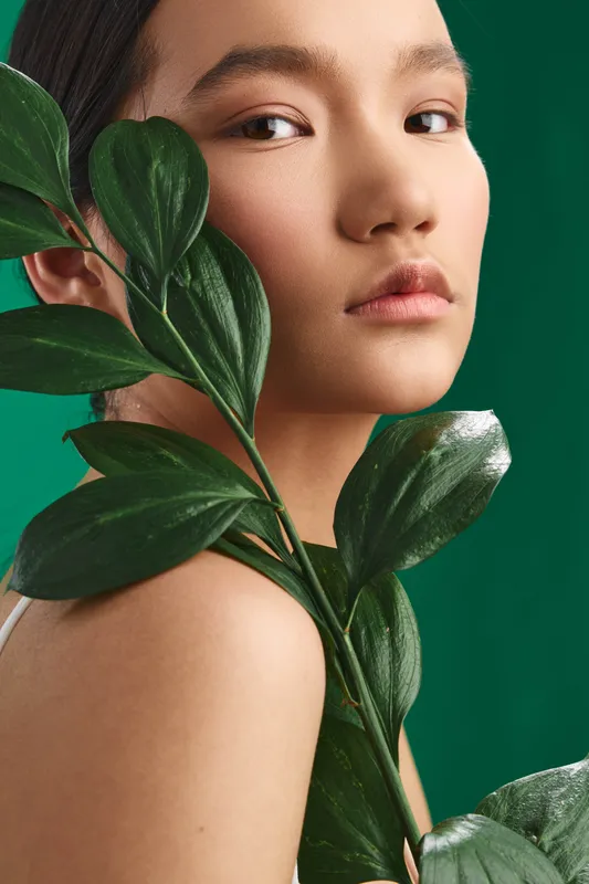 Asian woman portrait with lush leaves draped across her shoulder, set against a vivid emerald background, conveying elegance, calm, and natural beauty in a refined studio mood.