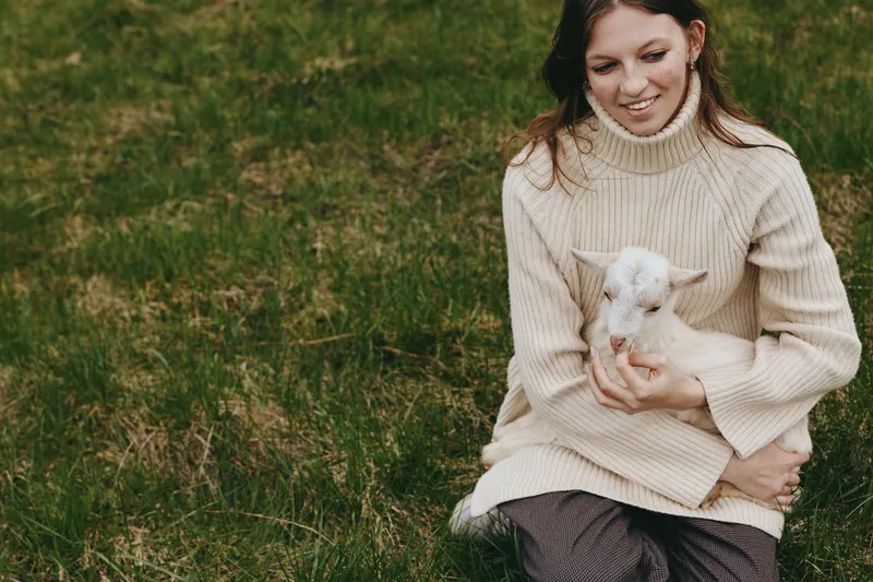 Cheerful woman outdoors, cradling a gentle kid goat in a cozy knit sweater, warm sunlight, relaxed pose, grassy park backdrop, natural colors, candid lifestyle moment, soft breeze, warm connection