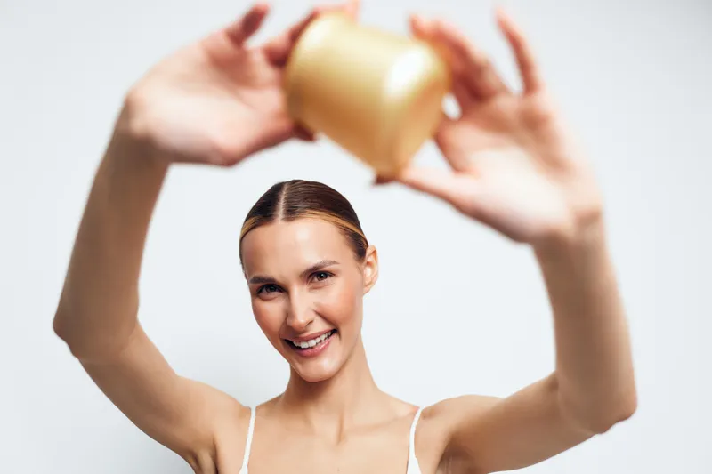 Smiling woman with clear skin holding cosmetic jar above head, bright studio background, natural beauty concept, skincare routine, youthful healthy appearance.