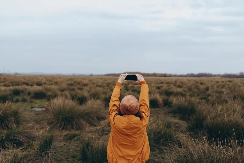 Woman outdoor field grassland lifestyle nature cloudy autumn calm portrait, standing in hooded orange coat on meadow, relaxed posture, serene horizon and cloudy sky, minimal background.