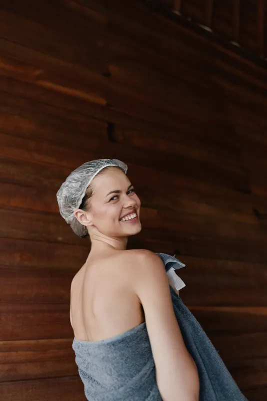 Smiling woman wrapped in towel with shower cap on head enjoying wooden sauna interior, conveying relaxation, wellness, and summer freshness in warm tones.