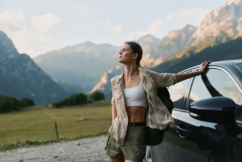 Young woman in casual outdoorswear leans on a car door in a mountainous landscape, enjoying fresh air, freedom, and a peaceful moment.