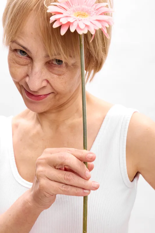 Woman portrait holding pink daisy flower on a stem, soft smile and thoughtful eyes, closeup against white background, gentle aging mood, calm expression