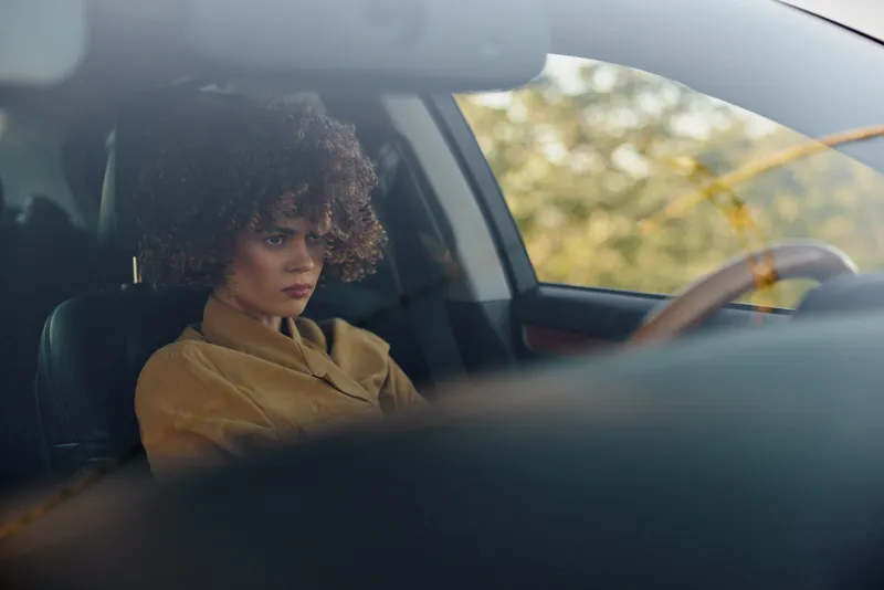A contemplative woman sits in the driver seat of a car, head bowed, in warm retro tones. Subtle burnout and introspection within a private, moody lifestyle moment.