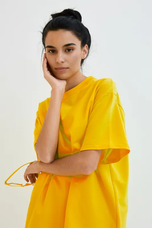 young woman wearing bright yellow oversized t-shirt with minimal makeup standing thoughtfully against plain light background in natural studio lighting