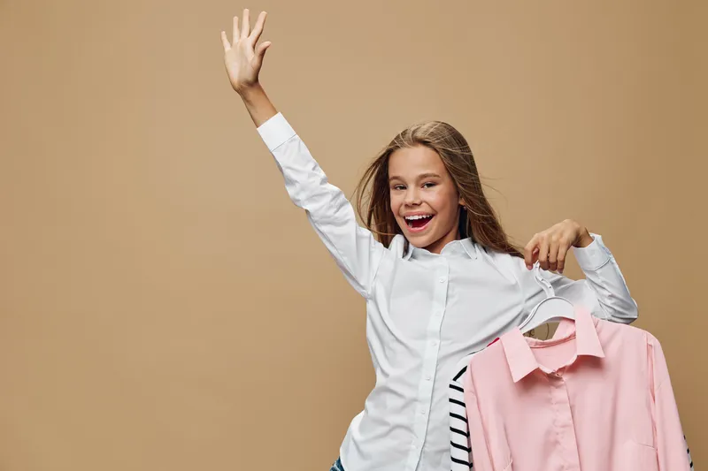 Happy girl waving hand and holding clothes on hangers smiling in casual outfit on beige background for shopping choice and style fashion concept.