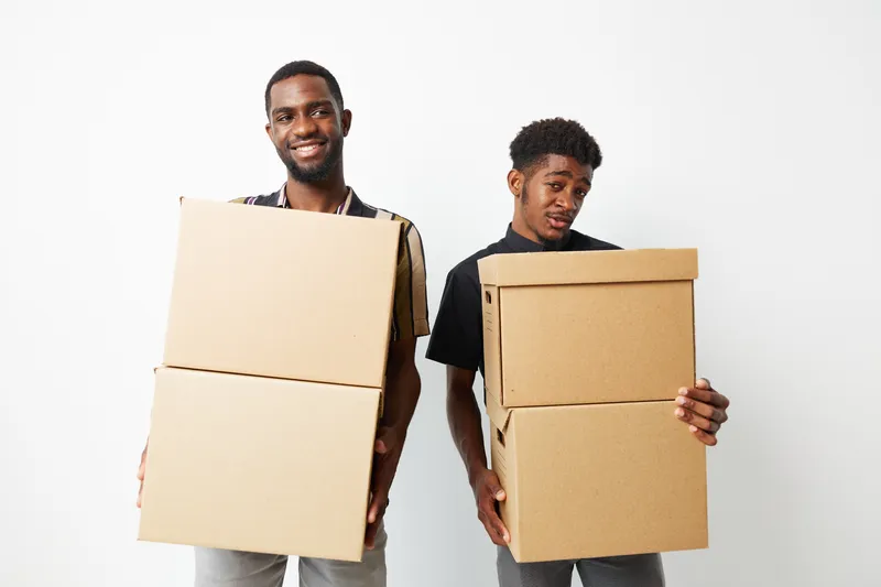 Two smiling African American men holding cardboard boxes in a bright, minimalistic setting. Their expressions show a mix of joy and playfulness, highlighting teamwork.