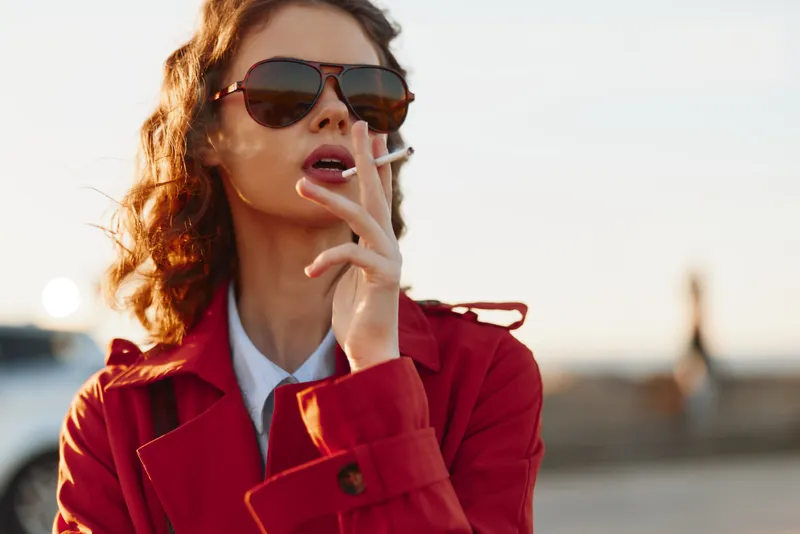 Young woman with curly hair wearing sunglasses and a red coat smokes a cigarette outdoors during golden hour with a thoughtful expression and soft natural light.