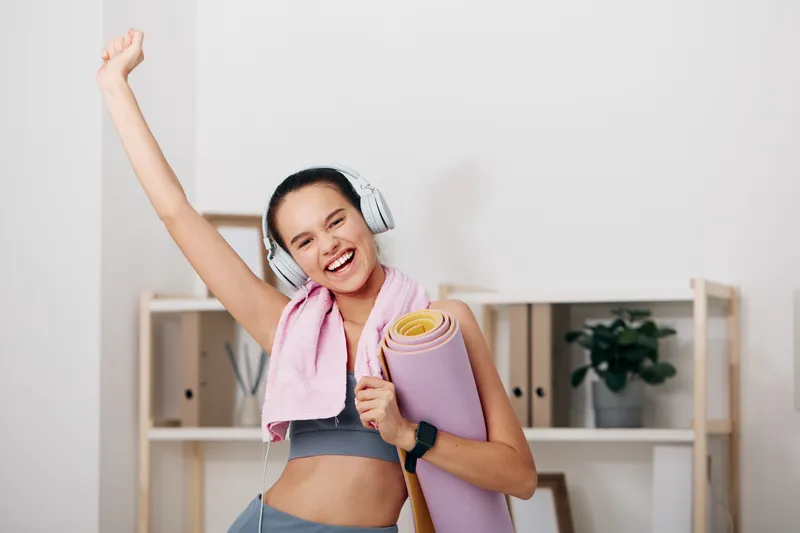 Woman fitness enthusiast joyfully raises a raised arm, holding a yoga mat and wearing headphones in a bright home studio, capturing a vibrant fitness moment.