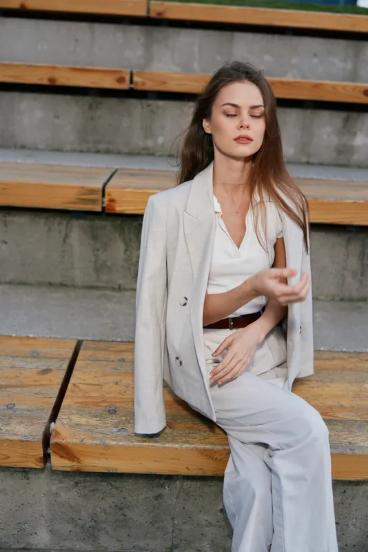 Woman in light beige suit sitting on wooden stairs with calm expression, natural makeup, casual business style, outdoor urban background, soft neutral tones