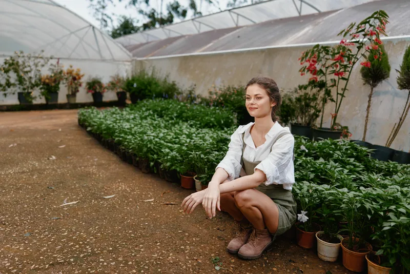 Young woman in garden kneeling among green plants, wearing casual attire and smiling, showcasing connection with nature and gardening passion