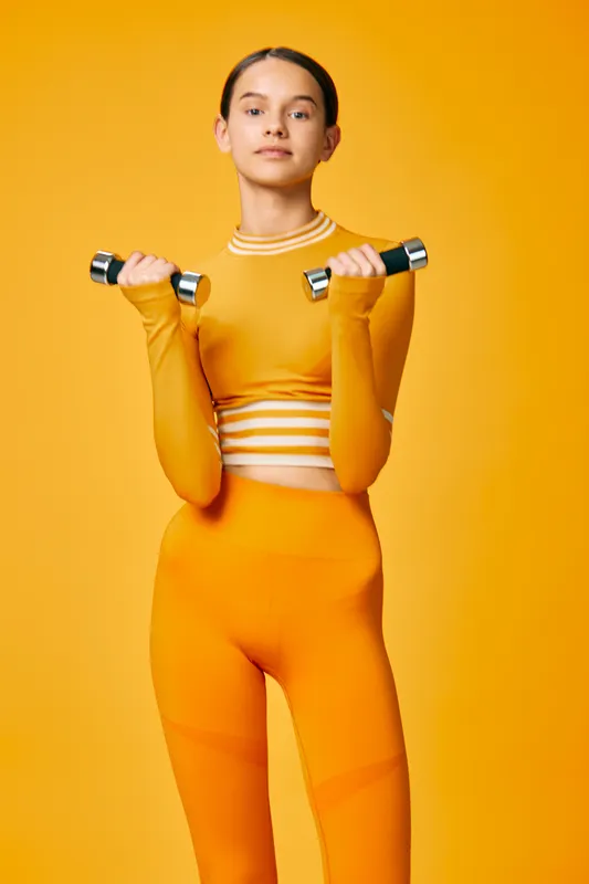 Fitness, woman and sport in a bold orange athletic set, lifting dumbbells in a studio. Dynamic workout scene with focused expression, highlighting strength and dedication.