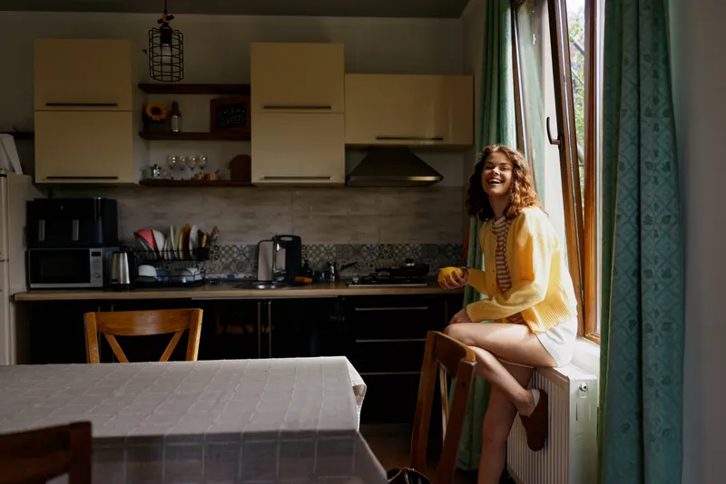 Smiling woman in a yellow cardigan sits on a kitchen windowsill, relaxed at home. Natural light, modern kitchen, casual lifestyle, cozy mood, everyday moment, cheerful vibe.