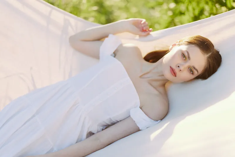 Young woman in a white off shoulder dress resting in hammock outdoors, exuding tranquility and serenity in a lush green environment