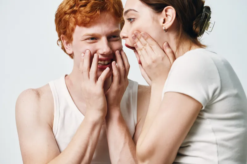 Young man and woman whispering secret with hands covering mouth in casual white clothing against light background in closeup gesture communication.