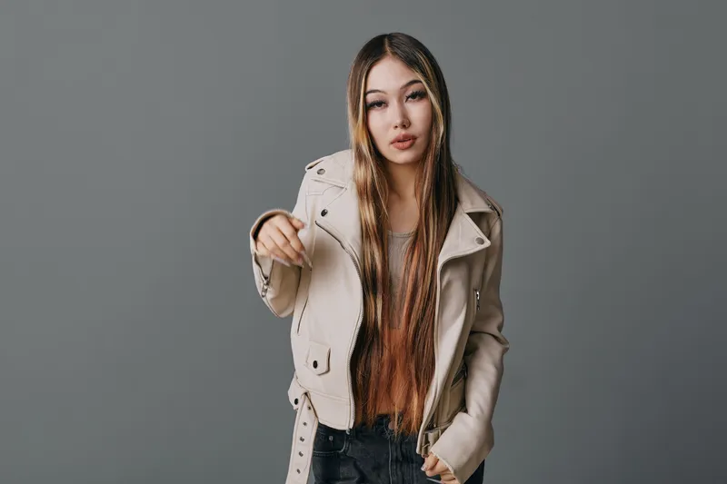 Young woman with long braided hair wearing beige leather jacket and casual clothes standing on gray background with confident expression pointing forward.
