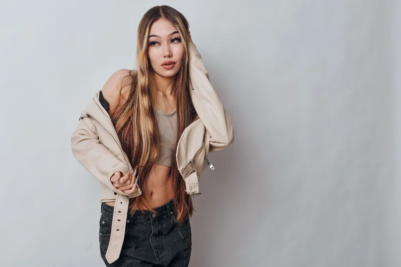 Young woman with long hair wearing beige jacket and crop top posing confidently on gray background with casual style and modern fashion look indoors.