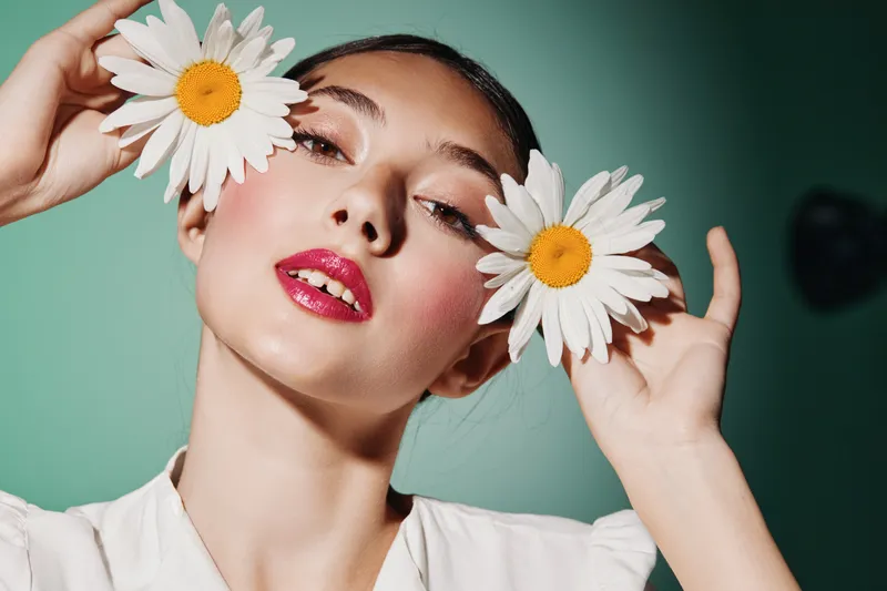 Young woman with bold red lipstick and shiny eye makeup holds two chamomile flowers near her eyes against turquoise background. Creative beauty and fashion concept.
