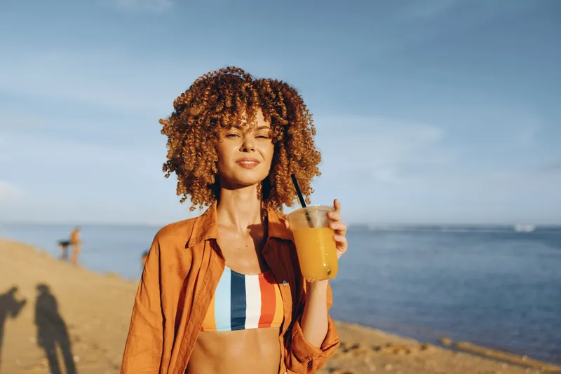 Woman at beach by the sea curly hair holding drink cup wearing an orange jacket and striped top relaxed smile summer lifestyle golden hour seaside travel portrait depth of field