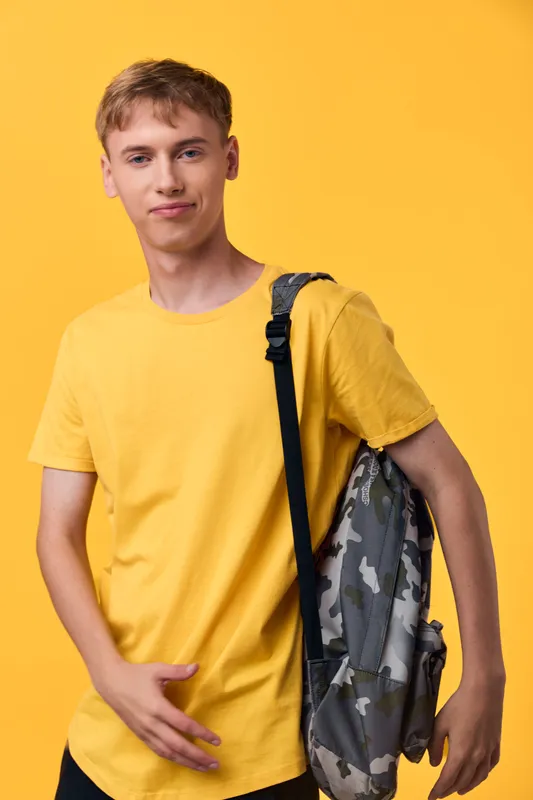 Student wearing a yellow t shirt with a camouflage backpack, posing against a bright yellow background, casual style and ready for class, travel, or daily adventures