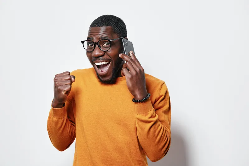 African American man with dark skin wearing glasses and orange sweater talking on smartphone with excited expression and fist pump gesture isolated on white background.