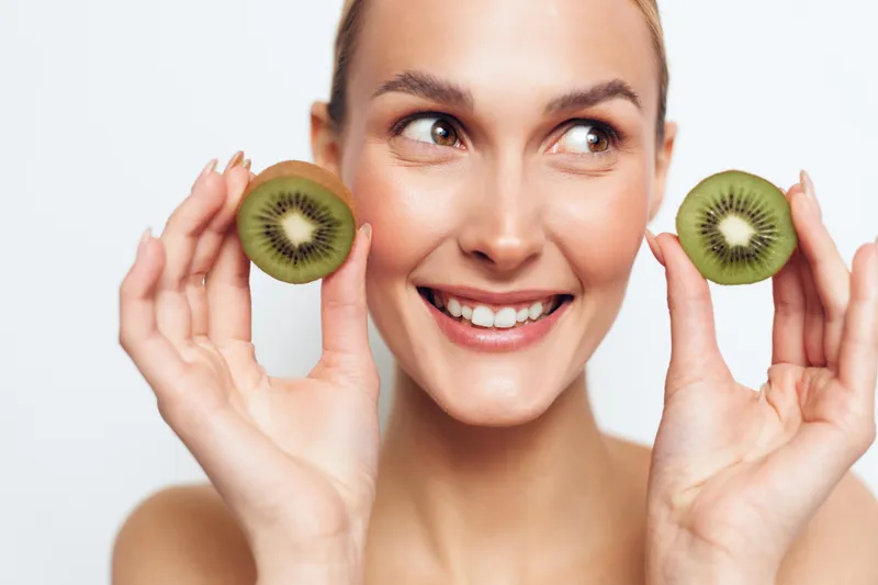 Smiling woman holds kiwi halves, showcasing healthy beauty and skincare, in a bright studio portrait with soft light, conveying freshness, cheer, and natural wellness