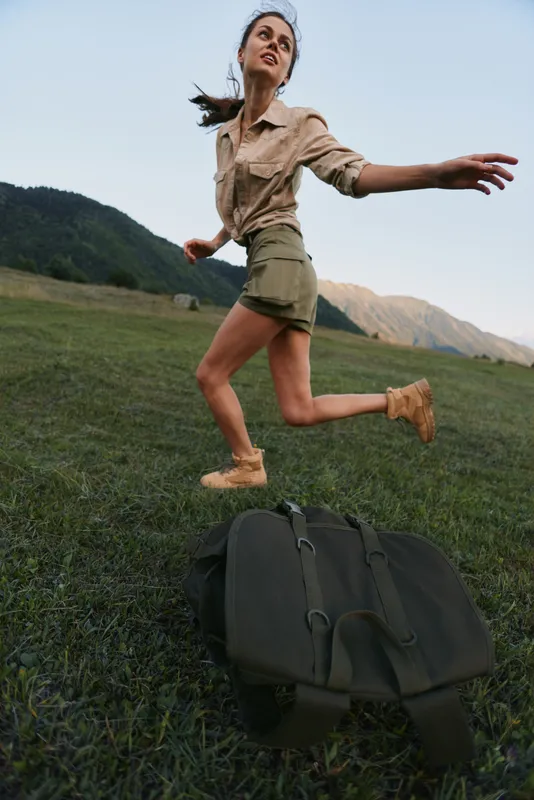 Woman in casual outfit leaps across a grassy meadow with a backpack, capturing carefree outdoor lifestyle and adventure amid mountain landscape.