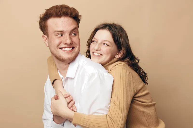 Happy young couple embraces and smiles looking at each other on beige background. Concept of love, relationship, friendship, and positive emotions between man and woman.