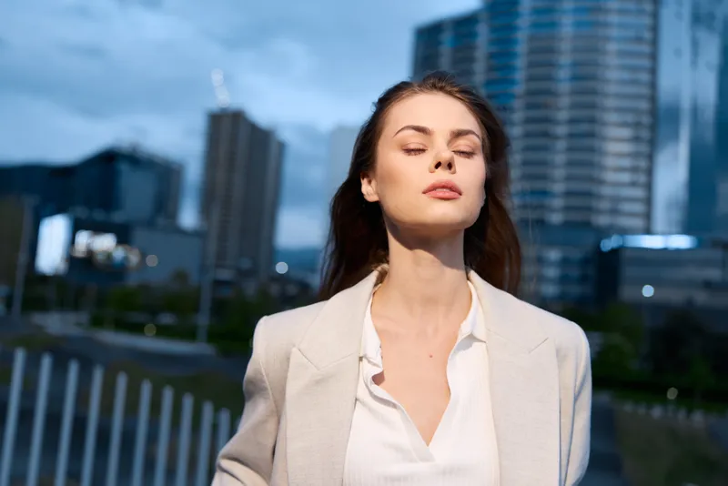 Confident businesswoman in urban setting, exuding professionalism and calmness, dressed in a light blazer, with a city skyline background at dusk.