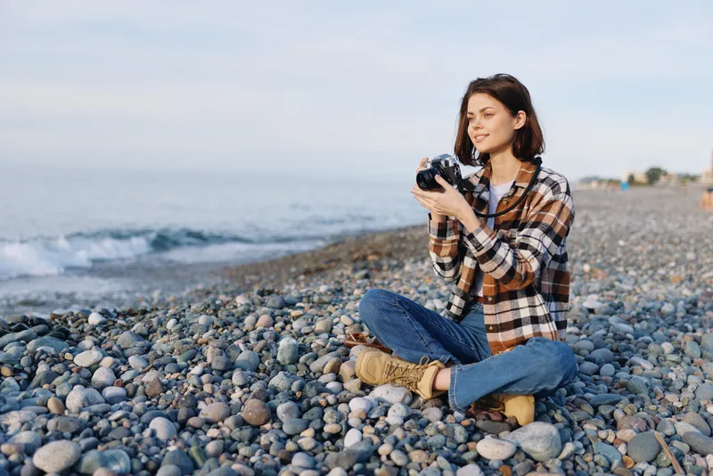 A photographer sits cross legged on a pebble beach by the sea, holding a camera, wearing a plaid shirt and jeans, focused and calm in natural coastal light.