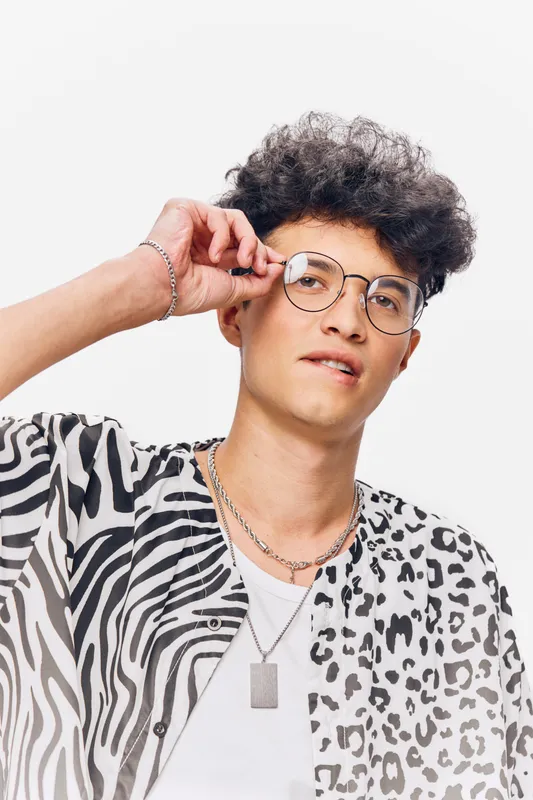 young man wearing round glasses adjusting them with hand, curly hair, stylish patterned shirt, casual look on white background