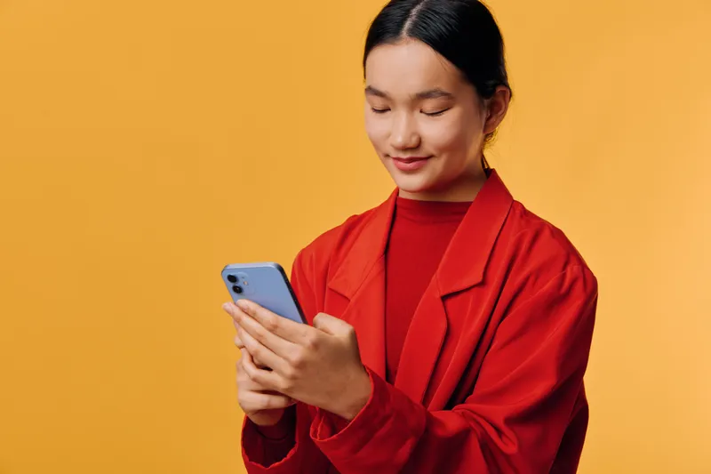 Young woman in red jacket using smartphone, smiling and looking at screen. Portrait of Asian girl with black hair, isolated on orange background, digital communication and modern technology concept.