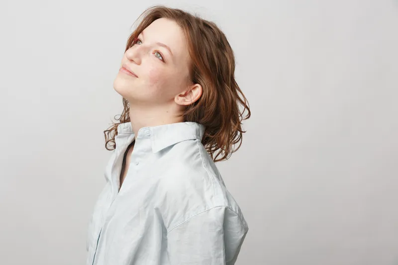 Young woman with curly hair wearing a white shirt looks up thoughtfully against a plain light background. Concept of hope, contemplation, and natural beauty in minimalistic style.
