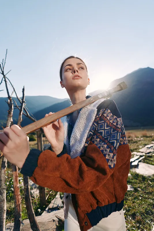 woman outdoors mountains hiking sweater sunlight portrait nature: Young woman in a patterned sweater jacket holds a wooden stick on a mountain trail, backlit by sun with a calm thoughtful expression.