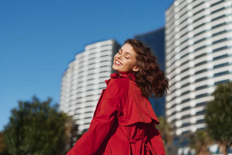 Happy young woman in red coat smiles with closed eyes enjoys sunny day outdoors near modern residential buildings with clear blue sky background.