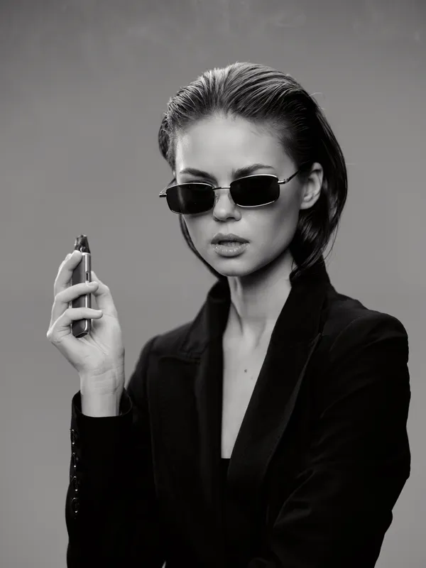 Black and white portrait of a stylish woman wearing sunglasses and holding a vape device, exuding confidence and modern elegance.