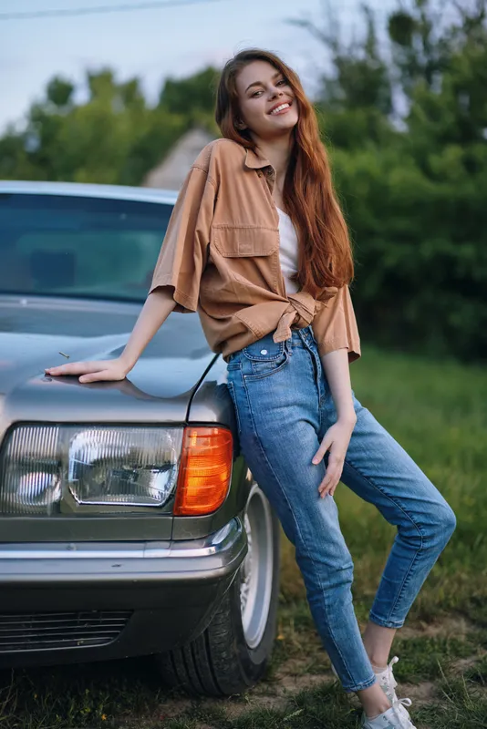 Young woman with long red hair in casual outfit leaning on vintage car in summer greenery, smiling with carefree emotions and relaxed demeanor.