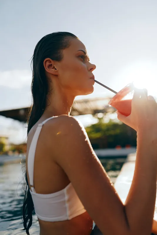 Cheerful woman outdoors, sipping a beverage through a straw by a sunny poolside, summer leisure and pool vibe, relaxed posture, warm light, vibrant reflection, casual lifestyle, refreshing moment