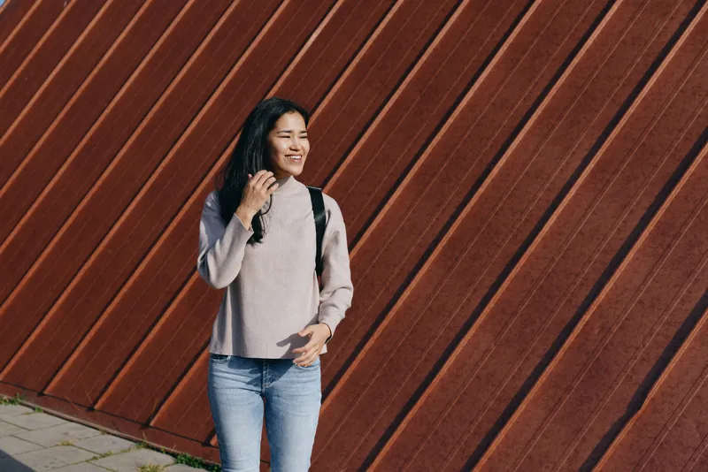 Woman with backpack, casual outdoor fashion sweater and jeans, smiling as she walks beside a diagonal wall, urban lifestyle scene with candid charm and everyday style, perfect for fashion, travel or