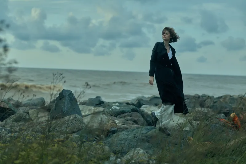 Woman walks along a rocky shore in a dark coat, windy weather, distant sea and cloudy sky, creating a moody, cinematic scene of solitude and exploration