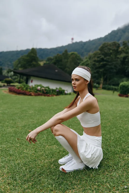 Young athletic woman in a white sports outfit squatting outdoors, showcasing fitness, health, and a vibrant green background in a summer setting