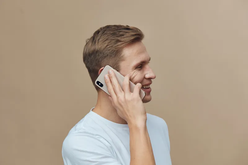 Smiling man talking on mobile phone with a cheerful expression, wearing a light blue T-shirt, indoor background, casual style, lifestyle concept, friendly communication.