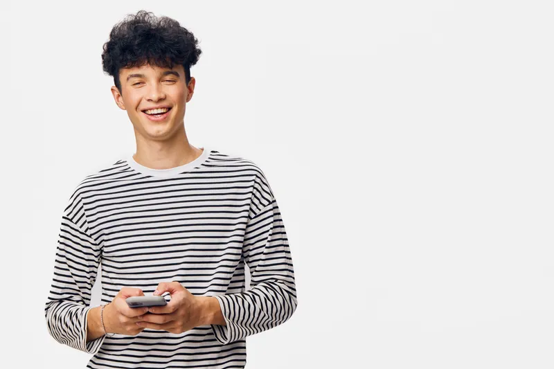 Smiling young man with curly hair wearing a casual striped shirt, holding a smartphone, expressing joy and natural happiness on a clean white background.