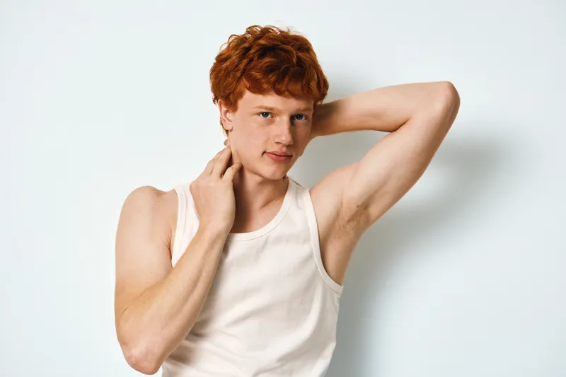 Young man with red hair wearing white tank top posing with hand behind head and another touching neck against white background, expression thoughtful and relaxed.