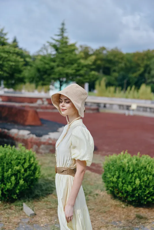 Elegant woman in a cream vintage dress and straw hat stands in a sunlit garden, relaxed pose, soft natural light creating a peaceful outdoor fashion scene.