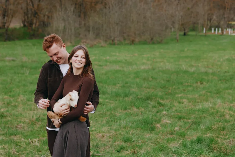 Cheerful couple outdoors, smiling with a small goat kid in a lush field, gentle breeze, warm clothing, relaxed pose, sunlit moment, natural colors, countryside backdrop, candid lifestyle scene